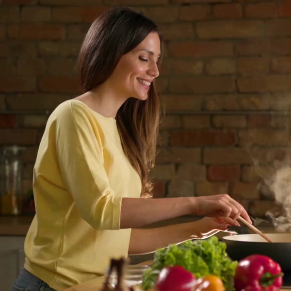A woman happily cooking with a pan and fresh vegetables.