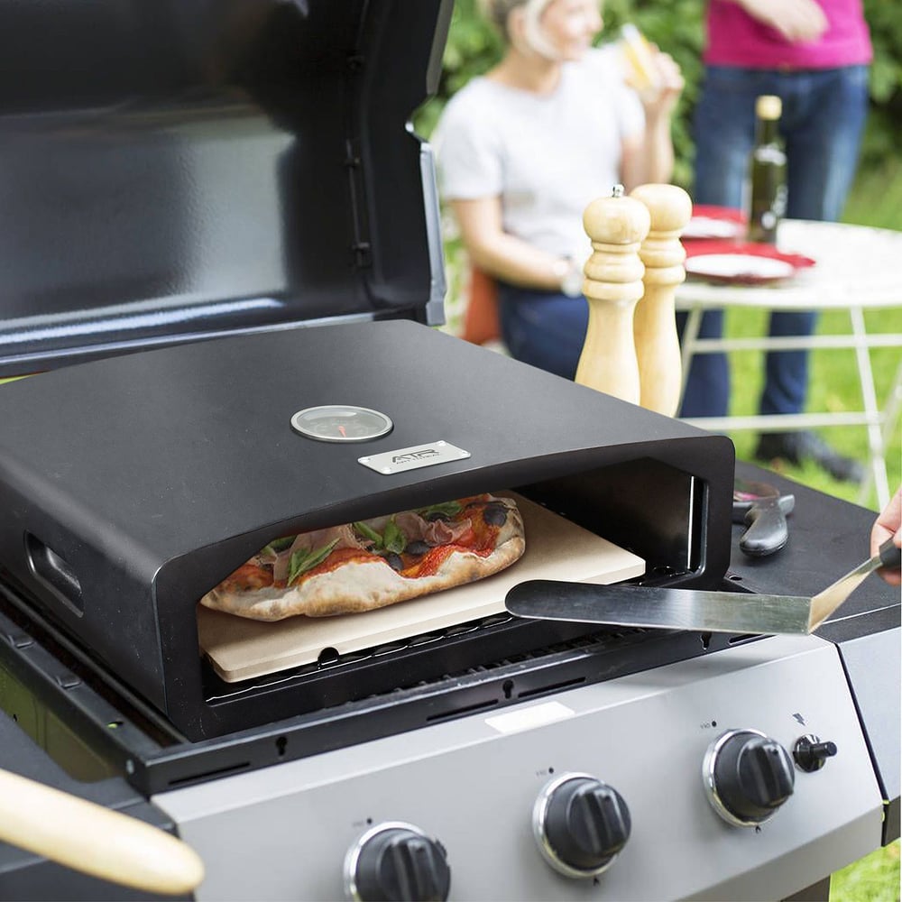 Outdoor pizza oven on a gas grill baking a fresh homemade pizza with people gathered in a garden setting.