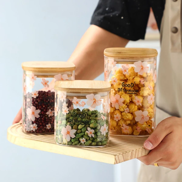 Person holding a tray with three decorative glass jars filled with green peas, red beans, and popcorn.