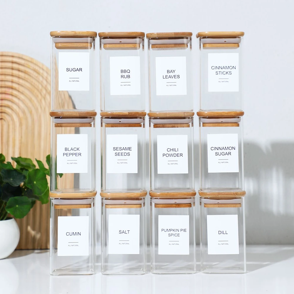 Glass spice jars in rows with wooden lids and labeled containers holding sugar, BBQ rub, bay leaves, cinnamon sticks, black pepper, sesame seeds, chili powder, cinnamon sugar, cumin, salt, pumpkin pie spice, and dill.