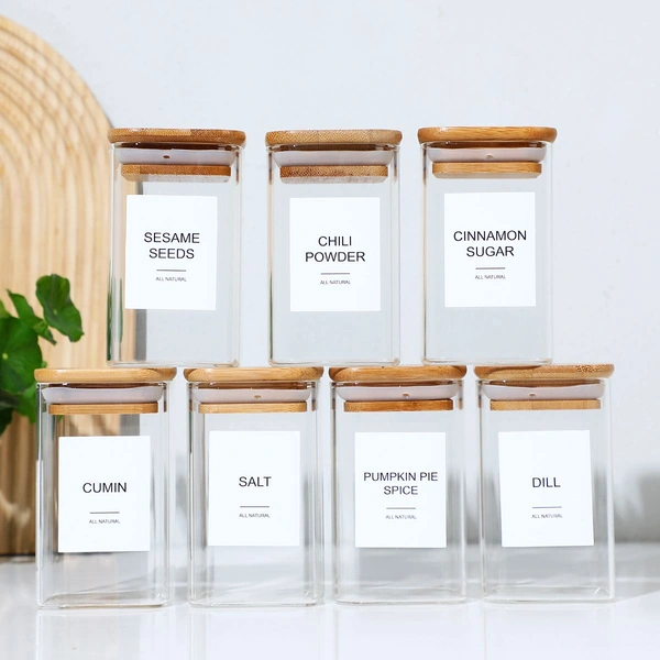Seven labeled glass spice jars with bamboo lids containing sesame seeds, chili powder, cinnamon sugar, cumin, salt, pumpkin pie spice, and dill.